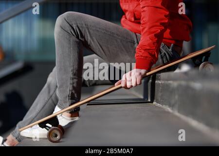 Stilvoller Mann, der in legerer Kleidung auf den Stufen sitzt, mit Longboard/Skateboard im Freien sitzt, Seitenansicht, zugeschnittenes Bild. Urban, Subkultur Stockfoto