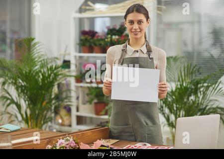 Taille hoch Porträt der lächelnden jungen Frau, die ein leeres weißes Schild hält, während sie im Blumenladen steht, Kopierraum Stockfoto