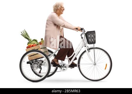 Profilaufnahme in voller Länge von einer älteren Dame, die auf einem Dreirad mit Obst und Gemüse in einer Kiste auf weißem Hintergrund reitet Stockfoto