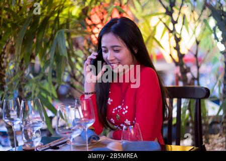 Eine junge Vietnamesin, die mit ihrem Handy ein traditionelles rotes Ao Dai Kleid trägt Stockfoto