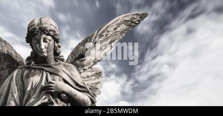 Traurige Wächterengelskulptur mit offenen Flügeln isoliert mit verschwommenen weißen Wolken dramatischer Himmel. Trauriger Engel trauriger Ausdruck Skulptur mit den Augen nach unten und Stockfoto