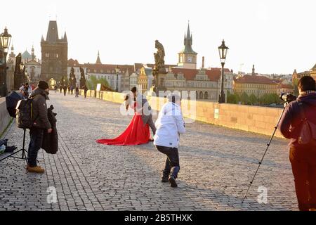 Asiatische Hochzeitsaufnahmen auf der Karlsbrücke Prag bei Tagesanbruch, wobei der Bräutigam die Braut in einem roten Kleid küsst, sanftes Frühlingslicht und ein dramatischer Moment Stockfoto