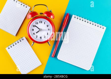 Retro-Wecker neben Stift und Notizbuch auf blauem und gelbem Hintergrund. Studium an Schule und Universität. Zeitmanagement Konzept, Business PLA Stockfoto