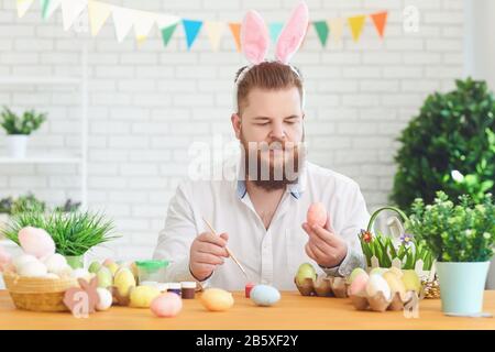 Frohe ostern. EIN lustiger fetter Mann schmückt Eier, während er an einem Tisch mit osterdekor im Hintergrund sitzt Stockfoto