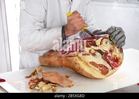 Metzger deboliert einen italienischen Schinken mit einem scharfen Messer in einer Metzgerei oder einem Geschäft in Nahsicht auf die Hände, eine in einem schützenden Eisenhandschuh Stockfoto