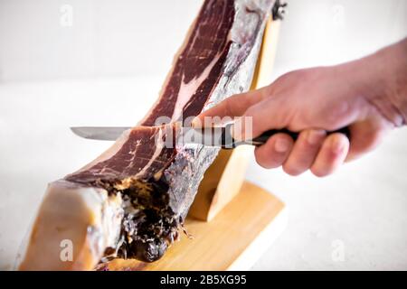 Nahaufnahme der männlichen Hand, die Jamon Iberica oder Pata Negra iberischen Schinken aus Spanien zuschneiden. Zugeschnittene Ansicht vor weißem Hintergrund Stockfoto