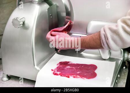 Metzger mit einer kreisförmigen Klinge, um rohe Rindersteaks zu schneiden, die sie sauber auf Papier darunter in einer Nahaufnahme an seinen Händen und der Maschine legen Stockfoto