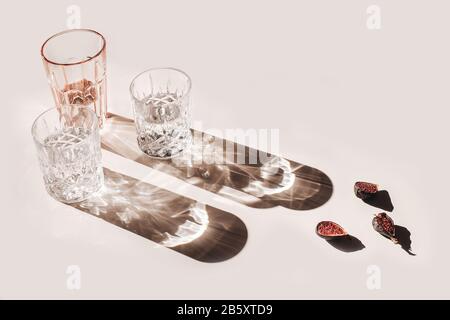 Sommer-Stillleben-Szene. Glitzernde Brille mit Wasser oder Cocktails und figs Obst auf pinkfarbenem Tischhintergrund bei Sonnenlicht schneiden. Lange, raue Schatten. Hoch Stockfoto