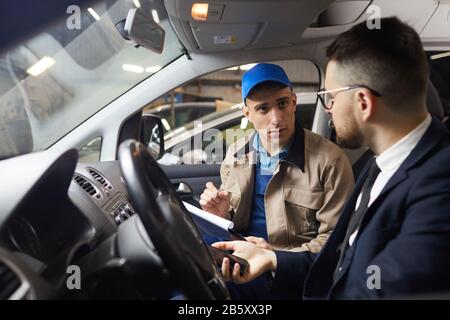 Horizontales Mittelporträt des professionellen Automechanikers, der mit seinem Kunden im Auto sitzt und ihm von Reparaturkosten und horizontalem Porträt erzählt Stockfoto