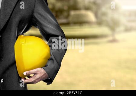 Geschäftsmann, der einen gelben Helm mit Baustellenhintergrund auf den Armen hält. Konzept des Arbeitstages Stockfoto