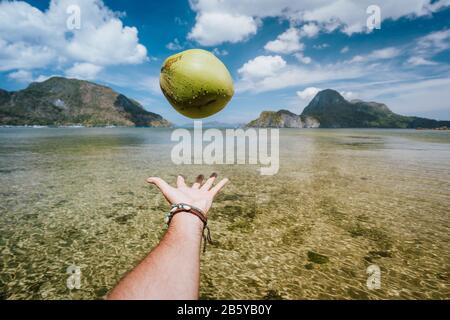 POV jongliert mit Kokosnuss in männlichen Händen gegen exotische Inseln in der Meeresbucht. Stockfoto