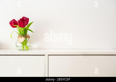 Rote Tulpen in hohem Glasbecher auf weißem Tisch gegen weiße Wand modernes helles Design, Federkonzept Stockfoto