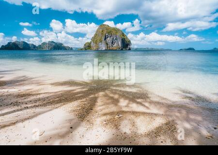 Palmen Schatten auf dem tropischen Strand Las Cabanas. Einzigartige tropische Insel im Hintergrund. Schöne Landschaft in El Nido, Palawan, Philippinen Stockfoto