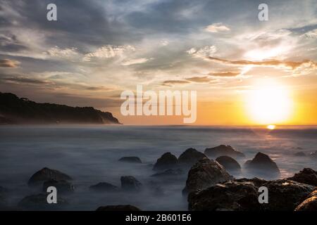 Atemberaubender Blick auf eine felsige Küste, die während des Sonnenuntergangs von einem glatten, seidigen Meer umspült wird. Melasti Beach mit seiner Klippe in der Ferne, Bali, Indonesien. Stockfoto