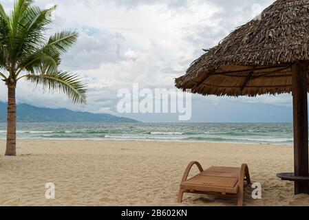 Urlaubsszene mit Liege- und Strohschirm am tropischen Strand gegen Meer und bewölkten Himmel. Mein Khe Strand in da Nang, Vietnam Stockfoto