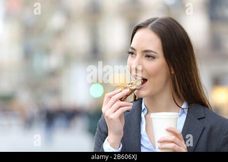 Portrait einer zufriedenen Geschäftsfrau, die eine Getreide-Snack-Bar mit einer Tasse Kaffee auf einer Straße in der Stadt isst Stockfoto