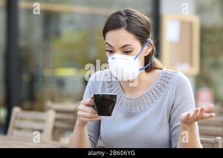 Verwirrtes Mädchen, das eine Tasse Kaffee mit einer Schutzmaske hält und dabei Ansteckungen auf einer Café-Terrasse vermeidet Stockfoto