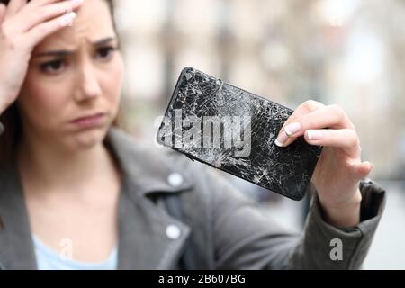 Nahaufnahme einer besorgten Frau, die ihr kaputtes Smartphone mit zerbrochenem Bildschirm auf der Straße hält Stockfoto