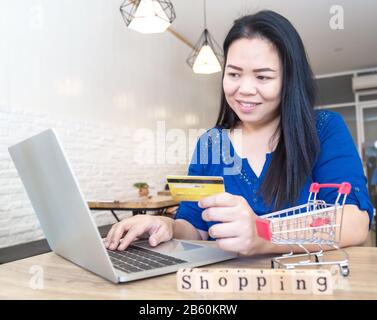 Frauen aus Asien haben eine Kreditkarte für Online-Shopping über den Laptop in einem Café. Auf einem Holztisch befindet sich ein aus Holzblo gefertigter Brief Stockfoto