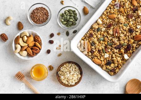 Hausgemachte Granola mit Nüssen und Samen auf Backblech mit Zutaten. Granola für gesundes Frühstück in Weiß, Draufsicht, flacher Lay. Stockfoto