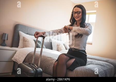 Eine glückliche Geschäftsfrau ist bereit, von Geschäftsreisen nach Hause zu gehen. Sie sitzt im Hotelzimmer und überprüft die Zeit auf der Armbanduhr. Stockfoto
