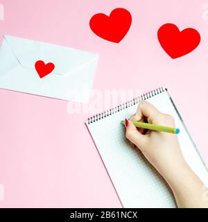 Mädchen schreibt einen Liebesbrief. Valentines Tageskonzept. Grußkarte zum valentinstag. Liebeserklärung. Einladung zur Hochzeit. Rote Herzen. Rosafarbener Hintergrund. Anzeigen Stockfoto