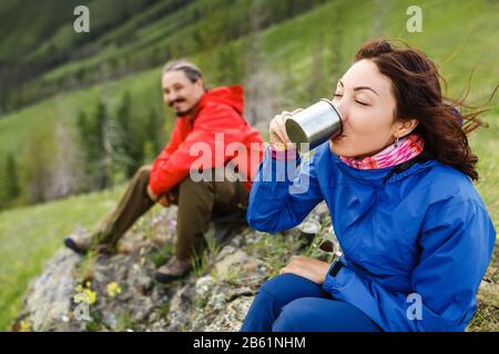 Seitenansicht des Wanderers fröhlicher junger Mann und Frau, die auf einem Gras auf der Spitze des Hügels sitzen und eine heiße Tasse Tee trinken Stockfoto