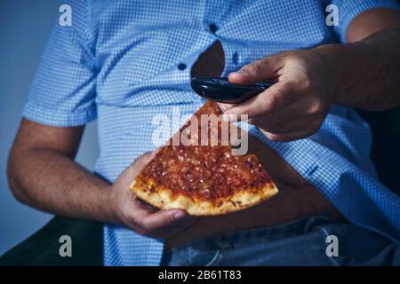 Nahaufnahme eines kaukasischen Mannes mit einem haarigen Bierbauch, auf dem Sofa sitzend, Pizza essen, während er den Fernseher beobachtet Stockfoto