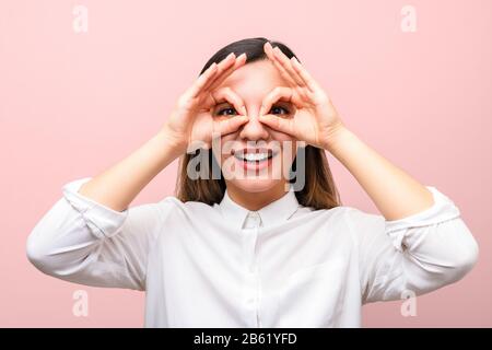 Attraktive Geschäftsfrau oder Studentin mit roten Lippen in weißem Hemd lächelnd und Brille aus ihren Fingern vor pinkfarbenem Hintergrund Stockfoto