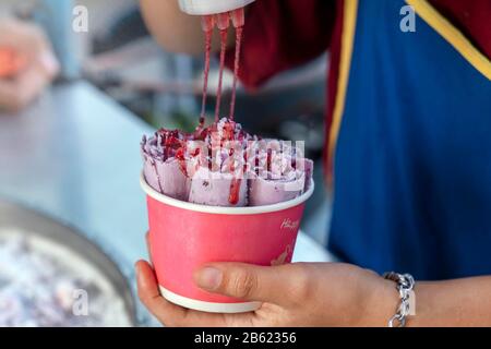 Die Zubereitung von Eiscreme, thailändisches Straßenessen. In einer Papierschale mit Kirschsirup violettes Eis kochen. Stockfoto