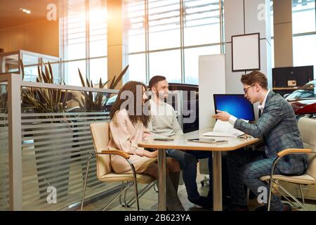 Junger, gutaussehender, eleganter Mann, der mit seinen Kunden, Arbeitszeiten und Arbeitsplatz am Tisch sitzt und die Merkmale des neuen Autos erläutert. Seitenansicht Foto Stockfoto