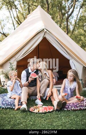 Fröhliche Familie, die Wassermelone beim Picknick auf der Wiese in der Nähe des Wigwam Tipi Zeltes isst. Mutter und Vater küssen, und zwei Töchter, die legere Kleidung tragen Stockfoto