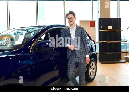 Mann steht vor einem Auto in einem Ausstellungsraum und posiert vor der Kamera, eleganter Händler schaut auf die Kamera. Nahaufnahme Foto. Kerl hat ein neues Auto gekauft Stockfoto