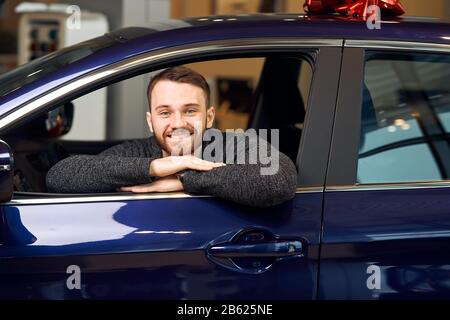 Bärtiger glücklicher Mann sitzt im neu gekauften blauen Auto mit roter Schleife auf der Oberseite und blickt auf Fenster im Autoshowroom.Glück, Schnäppchen, Träume werden wahr. C Stockfoto
