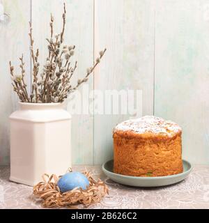 Osterzusammensetzung aus hausgemachtem Osterkuchen, blauem Ei im Papiernest und Weidenzweigen in weißer Vase auf Leinentischdecken an leicht bemalter Holzwand Stockfoto