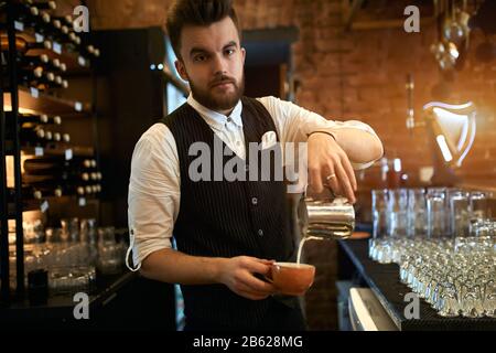 Professioneller, guter Barista, der die Milch in eine Tasse gießt, die Kamera betrachtet, im Coffeeshop auf die Kamera posiert, Photo.prepa aus der Nähe Stockfoto