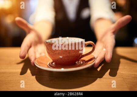 Barista in stilvollem Anzug, der eine Tasse frischen Kaffee serviert. Tasse Kaffee in den Händen des Kellners. Unvergesslicher Geschmack des besten Kaffees Stockfoto