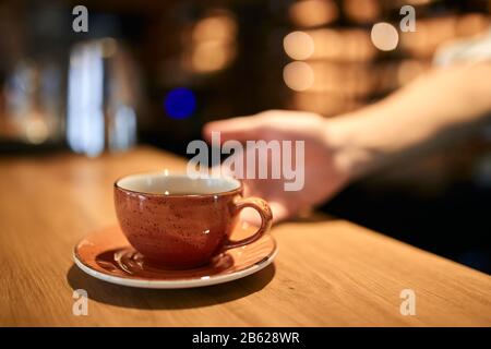 Nahaufnahme des Mannes, der Kaffee serviert, während er im Café steht. Konzentrieren Sie sich auf eine orangefarbene Tasse Tee, verschwommener Hintergrund. Stockfoto