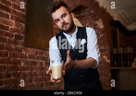 Servicemann in stilvollem Anzug, Weste und Hemd, das ein Glas Latte hält, Typ, der Kaffee für Kunden vorbereitet, Kunden im Café mit Loft-Interieur, Job, Stockfoto