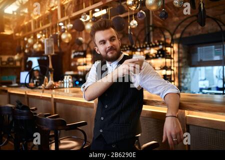 Ein glücklicher junger Mann, der ein Glas Latte hält, wird Freude daran haben, es zu verkosten, sich auf den Tisch zu lehnen und zur Seite zu schauen, und der Typ hat eine Ruhe bei der Arbeit Stockfoto