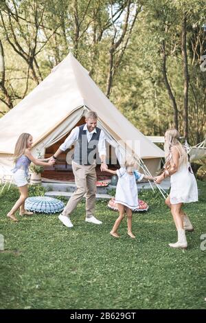 Junge, stilvolle Familie mit Kindern hat Spaß in einem Park. Vater, Mutter und Töchter tanzen in der Natur zusammen und halten die Hände. Familie tanzt in der Nähe Stockfoto