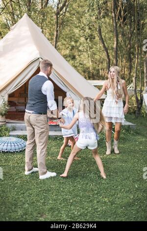 Junge Familie spielt an einem sonnigen Tag im Park zusammen. Vater, Mutter und zwei Töchter in legerer Boho-Kleidung, die tanzt und springt, halten die Hände in der Hand Stockfoto