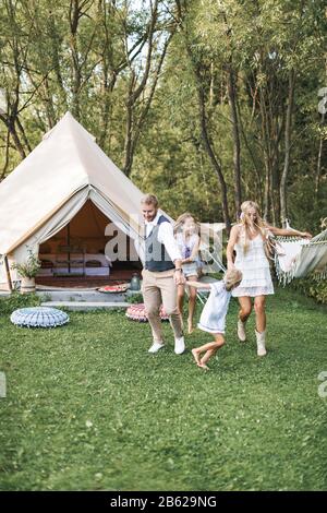 Glückliche Familie: Mutter, Vater, zwei Kinder Töchter auf der Natur, tanzen und laufen zusammen. Fröhliche Familie in boho-leichter Kleidung Stockfoto