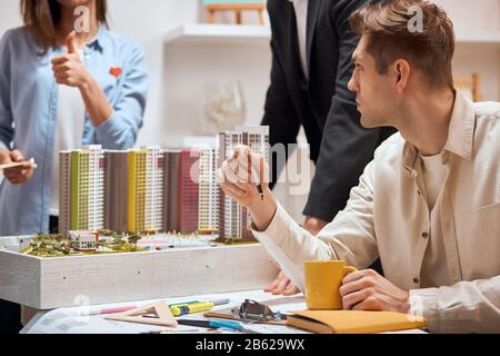 Fleißige junge Leute, die Architektur machen kleine Modell-Dummy-Bauarbeiten. Nahaufnahme Foto.junger blonder Mann sitzt mit einem Becher am Schreibtisch, während sein Stockfoto