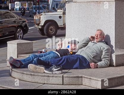 London, Westminster. Zwei Männer, die sich bei Sonnenschein auf dem Sockel der Edith-Cavell-Gedenkstätte am St. Martin's Place entspannen. Stockfoto