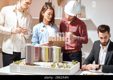 Talentierter Architekt im Bereich des roten Hemdes, der seine Kollegen über den zukünftigen Bauplan berät. Nahaufnahme Foto. Sekretärin, die einen Ordner hält, in dem sie ihren Vorgesetzten que fragt Stockfoto