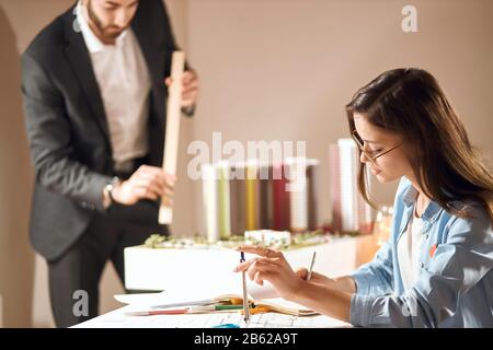 Junge Frau und Mann, die an Blaupause arbeiten, Nahaufnahme eines 3/4-Seitenbildes. Mann in elegantem Anzug, der einen Lineal im verschwommenen Hintergrund hält Stockfoto