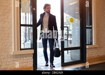 Junger Geschäftsmann, der eine schwarze Ledertasche hält und die Tür des Büros öffnet, Foto in voller Länge. Seltsamer Mann, der über Raub nachdenkt Stockfoto