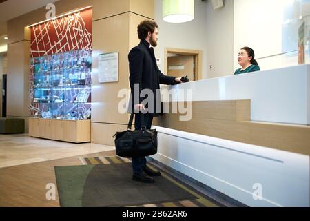 Ein junger, gutaussehender Geschäftsmann mit abag in den Händen, der mit weiblicher Rezeptionistin flirtet, ein Foto mit seitlichem Blick in voller Länge. Mann, der ein Zimmer gebucht hat, Tisch Stockfoto