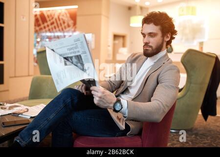 Junger cleverer Mann, der Zeit damit verbringt, die Zeitung im Saal des Hotels zu lesen, Nahsichtfoto Stockfoto